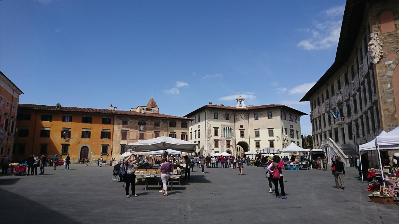 A local market on Piazza dei Cavalieri, Knights' Square, with tourists growing the stalls and Palazzo della Carovana on the side on a sunny spring day