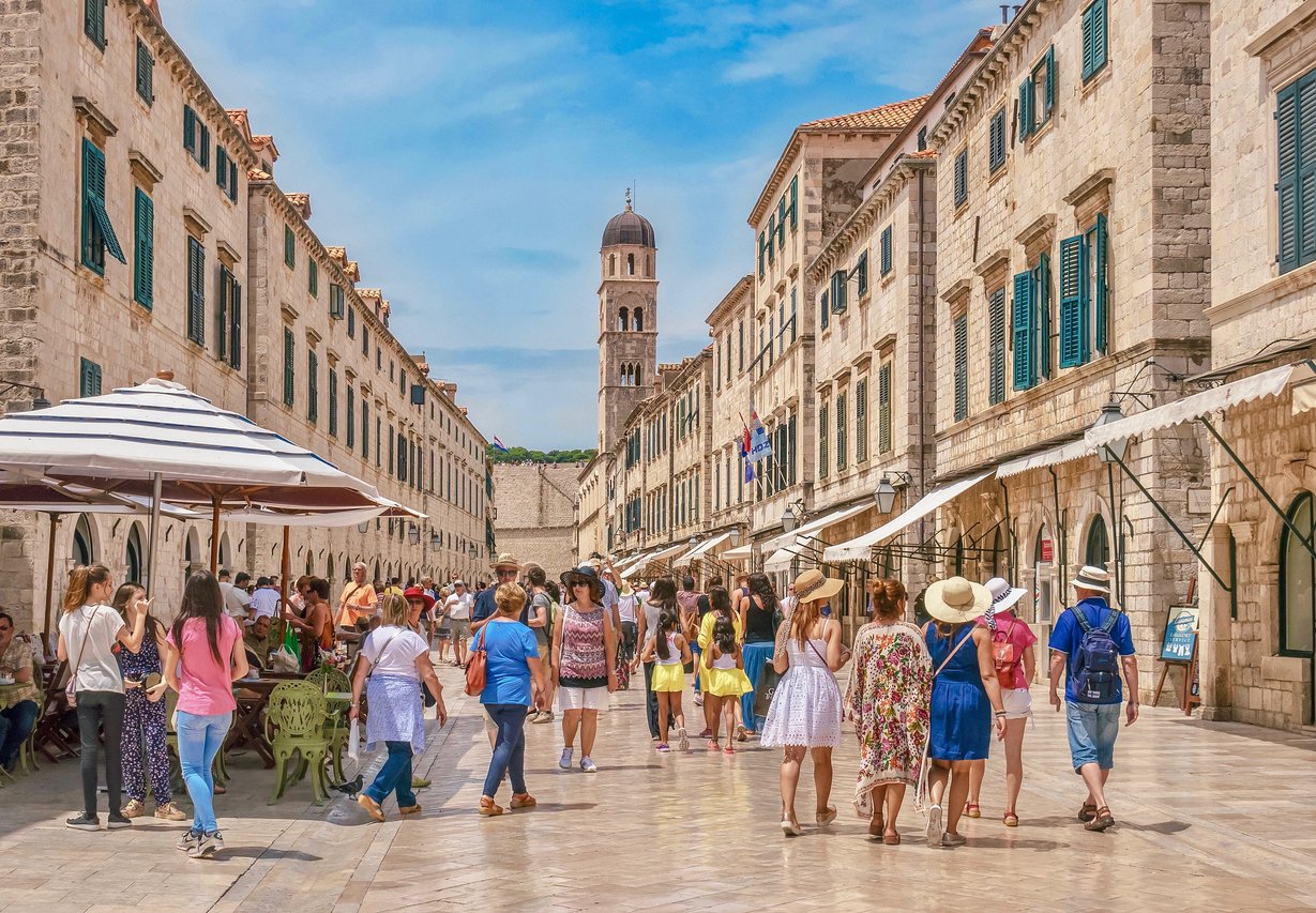 Tourists wearing summer clothing walk along the main street of old Dubrovnik, looking at the shops, restaurants and limestone buildings.